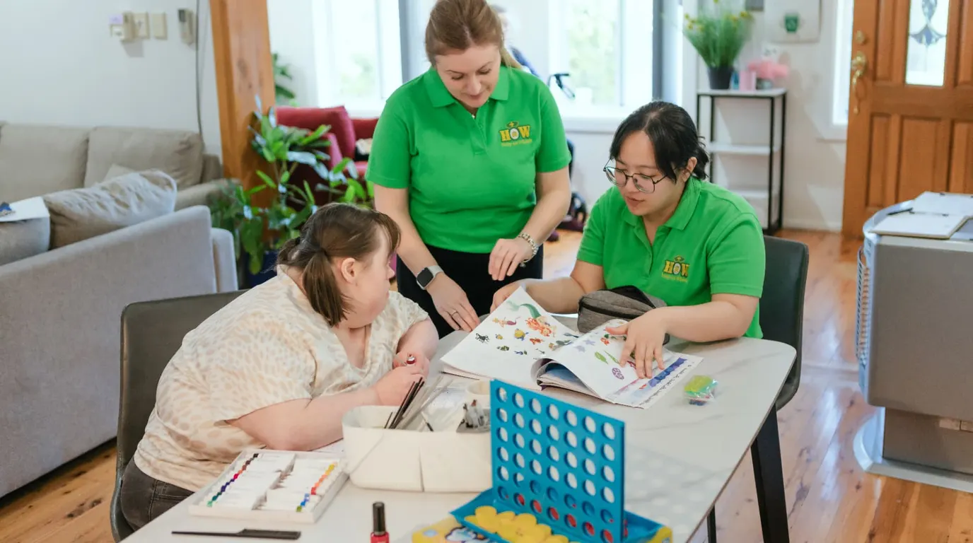 Two Help on Wheels support workers doing craft and games at a table with a young woman at home
