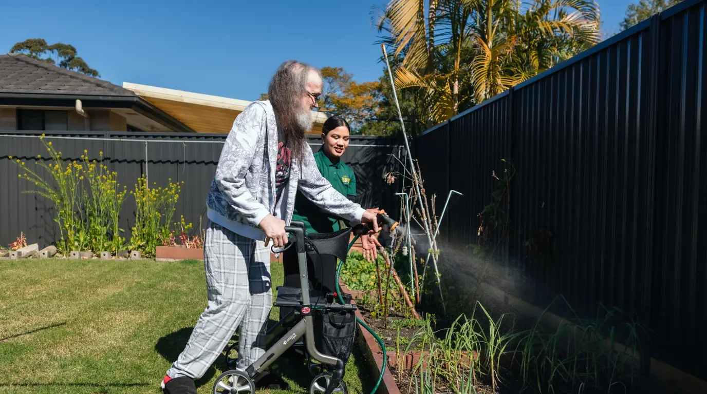 A Help on Wheels support worker helping a man with a walking frame water the vegetable garden in his backyard