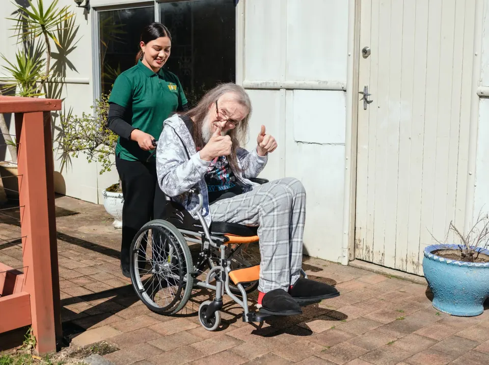 A Help on Wheels support worker taking a man in a wheelchair outside as he gives a thumbs up