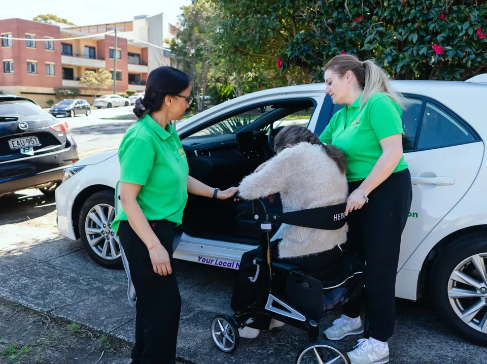 Two Help on Wheels support workers helping a participant with a walker into the car